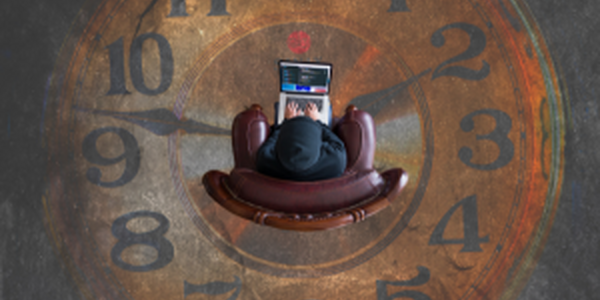 Person in dark hoodie sitting in chair and working on laptop with a clock prominent in background