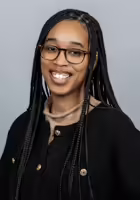Rainah Folk's headshot. Folk is wearing dark brown glasses and a black cardigan with a light brown mock neck sweater. 